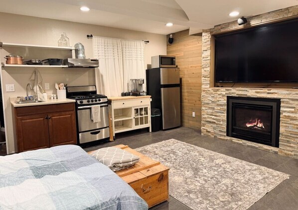 A compact living space with a kitchenette, a bed, a fireplace, and a TV mounted on the wall.