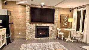 A living room with a fireplace, a flat-screen TV mounted above it, a dining area with a table and chairs, and a kitchen area with appliances.