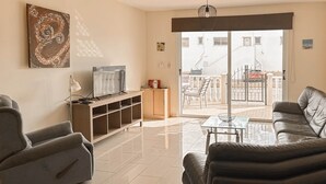 Sala de estar com sofá, televisão, mesa de centro de vidro e vista para o exterior.