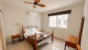 Quarto com cama, mesas de cabeceira, escrivaninha, cadeira, ventilador de teto e janela.