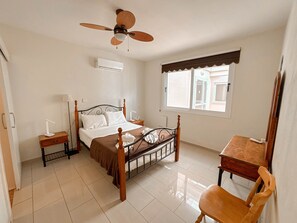 Un dormitorio con cama, mesitas de noche, escritorio, silla, ventilador de techo y ventana.
