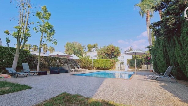 Piscina com espreguiçadeiras, área de estar coberta e um jardim bem cuidado com árvores e sebes.