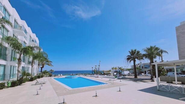 A swimming pool area with a clear view of the ocean, surrounded by palm trees and modern buildings. - Coralli Privileges Apt (Protaras)