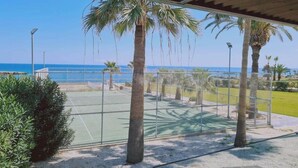 A tennis court with a net, surrounded by palm trees and a view of the sea. - Coralli Privileges Apt (Protaras)