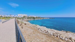 A coastal road with a paved walkway, rocky shoreline, and buildings in the background. - Coralli Privileges Apt (Protaras)