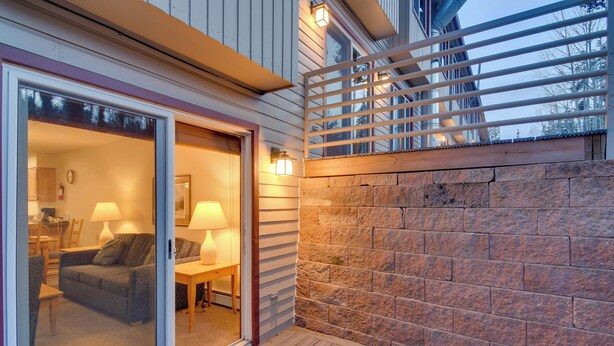 A modern house with a sliding glass door leading to a living room with a sofa, table, and lamps. There is a stone wall and a wooden deck. - Shuttle to Winter Park Resort! Group Condo w/ Pool (Winter Park)