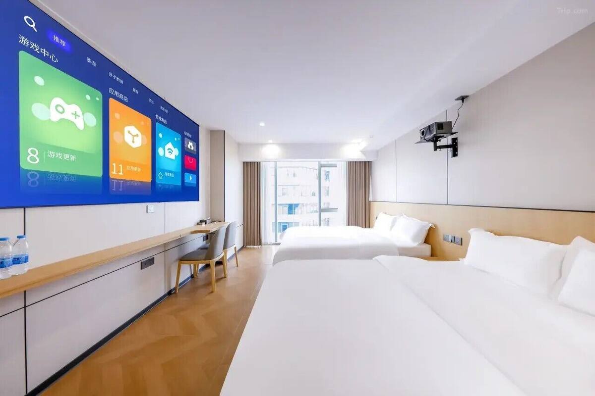 A hotel room with a large digital display on the wall, a bed, a desk with a chair, and a view of the outside through the window.