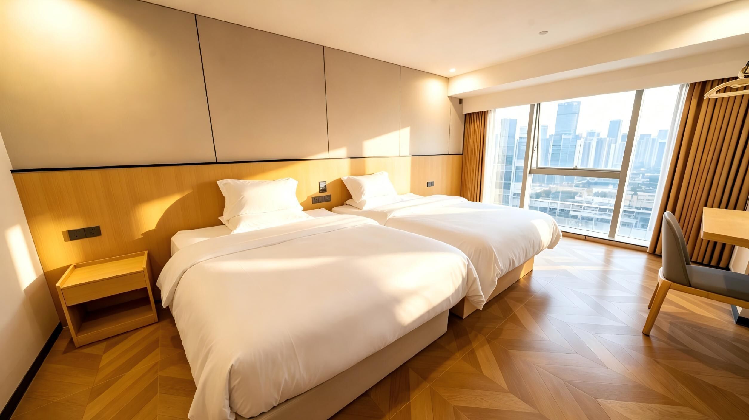 A hotel room with two beds, a wooden floor, a city view, and a desk with a chair.