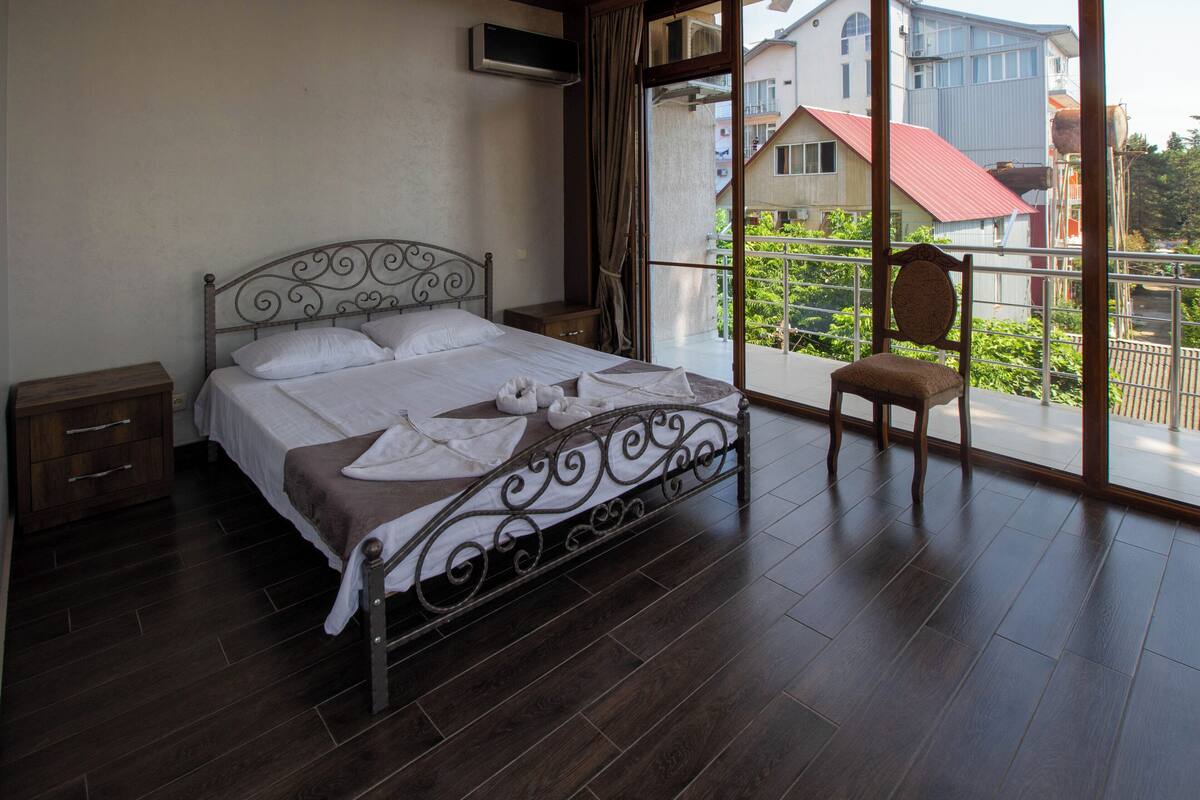 A bedroom with a metal bed frame, a wooden nightstand, a chair, and a view of buildings through a large window.
