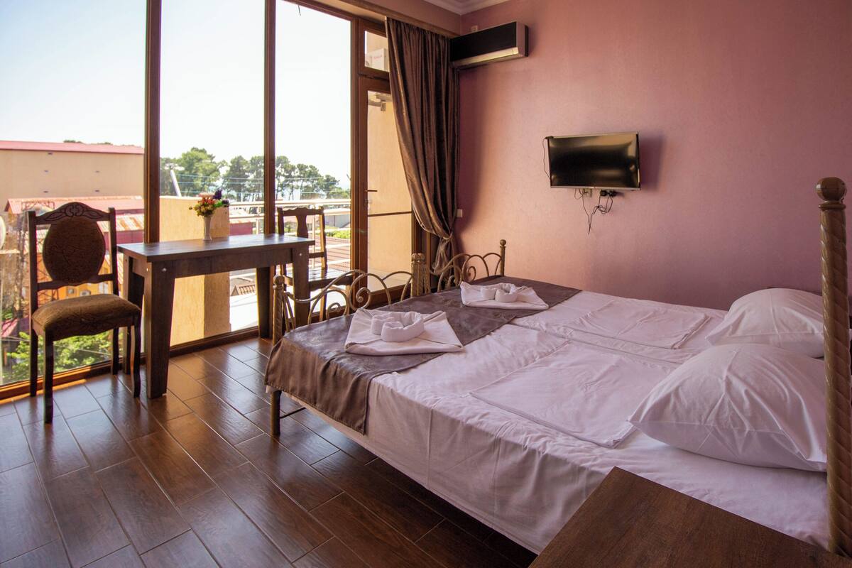 A hotel room with a bed, a desk, and a chair. There is a TV on the wall and a window with curtains.