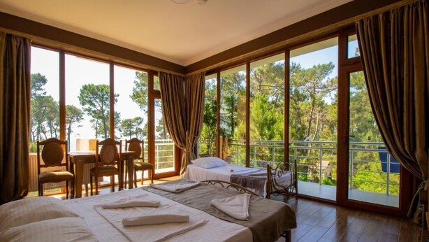 A bedroom with two beds, a balcony with a table and chairs, and large windows offering a view of trees. - Inter (Ureki)