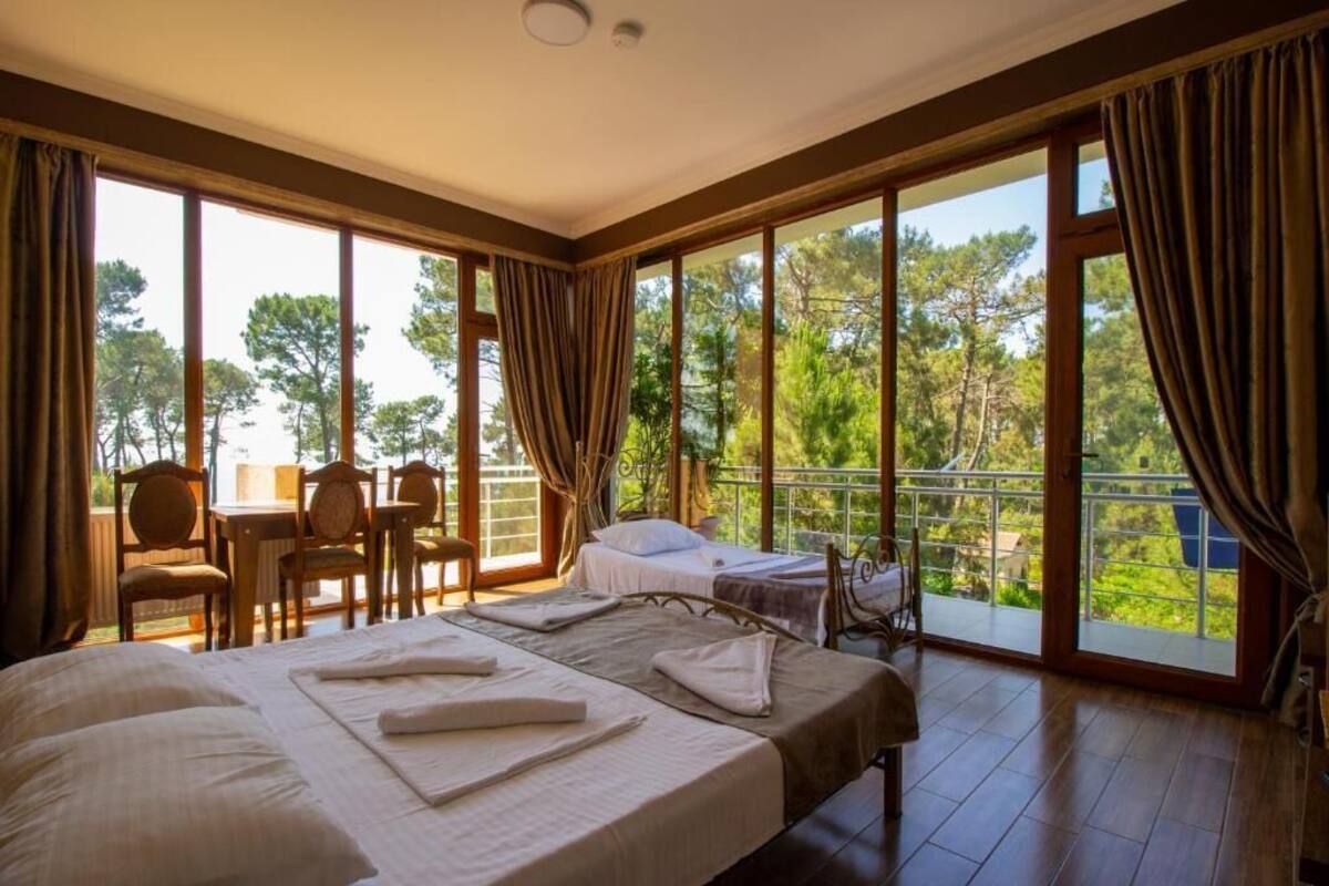 A bedroom with two beds, a balcony with a table and chairs, and large windows offering a view of trees.