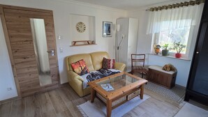 Een woonkamer met een bank, een bijzettafeltje, een houten stoel, een klein tafeltje met een lamp, een klok en een raam met planten.
