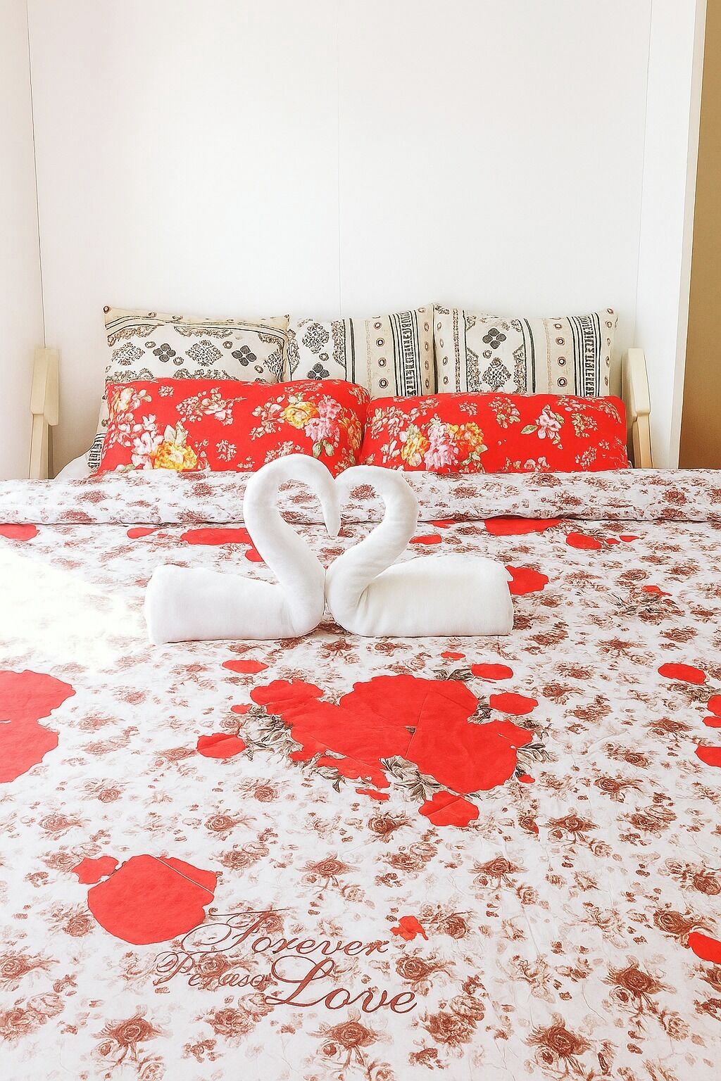 A bed with a floral bedspread and heart-shaped pillows, a heart-shaped towel arrangement, and a pillow with the text "Forever, Passion, Love".