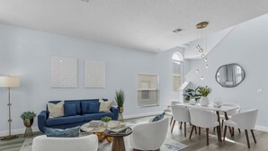 A modern living room with a blue sofa, white chairs, a coffee table, and a dining area with a white table and chairs. - Spacious 6-bedroom house with Private Pool/Spa, Game Room, & Resort Perks (Clermont)