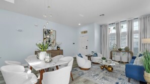 A modern living room with a dining area, featuring a white table, white chairs, a blue sofa, and a marble-patterned rug. - Spacious 6-bedroom house with Private Pool/Spa, Game Room, & Resort Perks (Clermont)