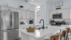 A modern kitchen with white cabinets, stainless steel appliances, a central island with chairs, and a coffee maker. - Spacious 6-bedroom house with Private Pool/Spa, Game Room, & Resort Perks (Clermont)
