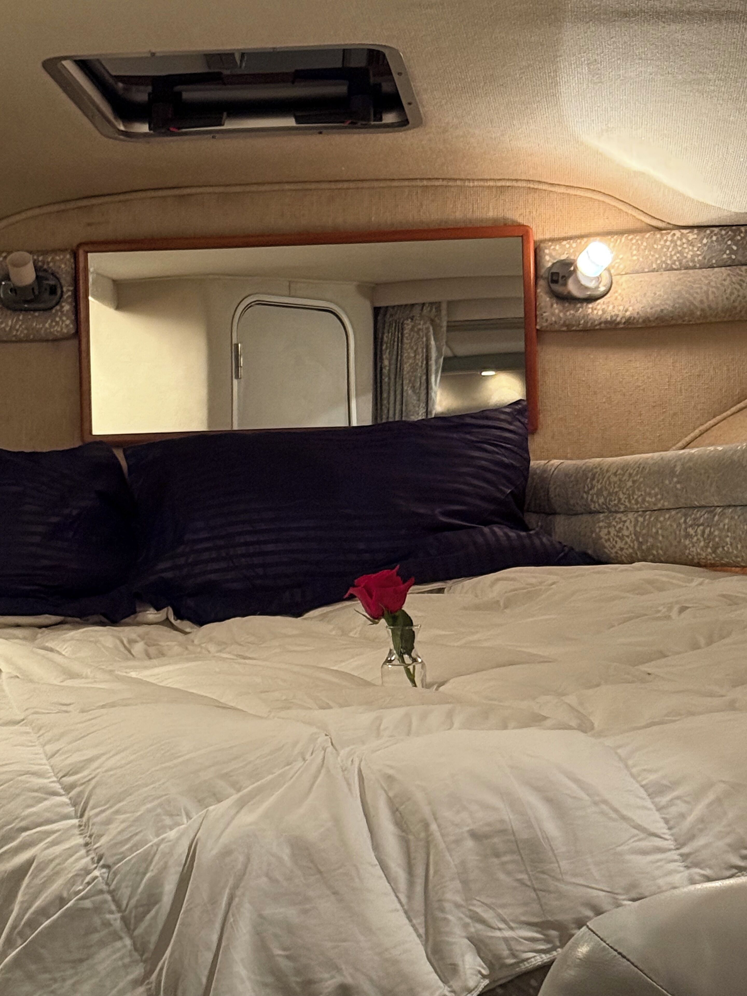A bed with a white comforter, a dark blue pillow, and a single red rose in a clear glass vase.