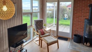 Ett mysigt vardagsrum med en tv, en bekväm fåtölj, en öppen spis och stora fönster med utsikt över omgivningen.