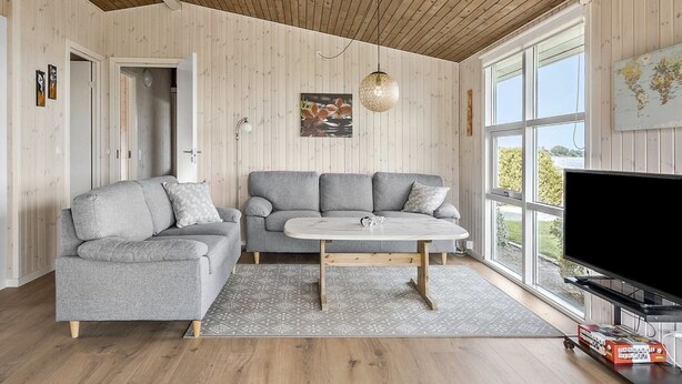 Ett modernt vardagsrum med ett träpanelat tak, två grå soffor, ett runt soffbord och ett stort fönster med utsikt över grönområden.