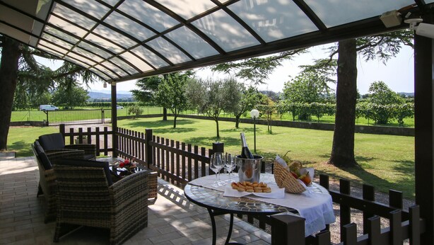 A covered outdoor seating area with wicker furniture and a table set for a meal, overlooking a grassy landscape with trees and a distant view of mountains. - Amazing home in Cividale del Friuli (Cividale del Friuli)