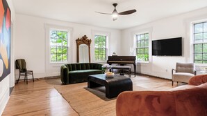 Un vaste salon doté de parquet, d’un canapé vert, d’un pouf noir, d’un piano et d’un téléviseur fixé au mur.