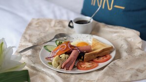 Ein Frühstücksteller mit Toast, Eiern, Tomaten und Gemüse auf einem Tisch. Im Hintergrund sind ein Bett und Kissen zu sehen.