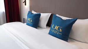 Ein Hotelzimmer mit einem großen Bett, zwei blauen Kissen mit dem 'UG Inn'-Logo, weißer Bettwäsche und einem roten Vorhang.