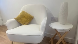 Een witte fauteuil met een gouden kussen, een klein wit tafeltje met een vaas en een houten vloer.