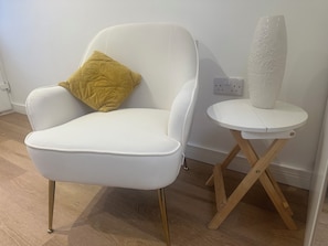 A white armchair with a gold cushion, a small white table with a vase, and a wooden floor. - Stylish studio with wet room & kitchen (London)