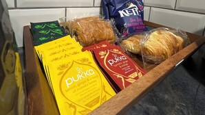 Een dienblad met diverse verpakte etenswaren, zoals snacks en brood, in een keuken met witte betegelde muren.