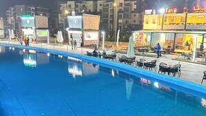 Zona de piscina nocturna con mobiliario exterior y letreros iluminados para restaurantes.