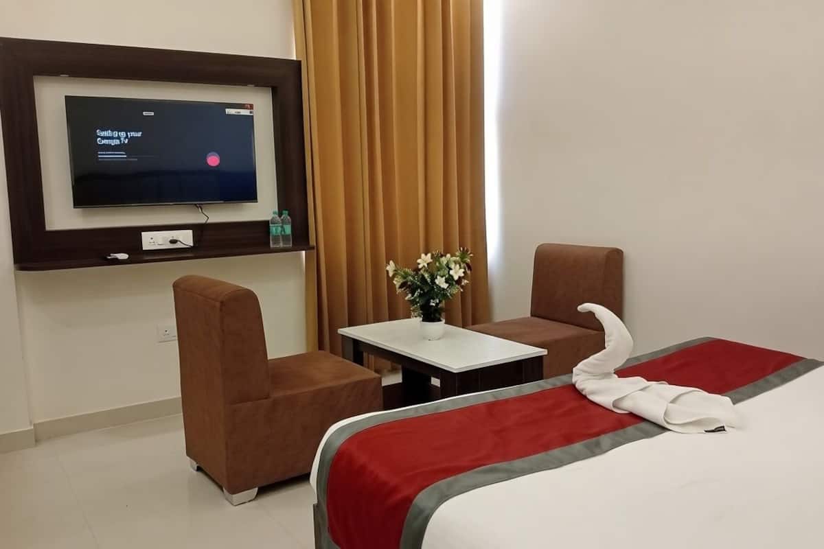 A hotel room with a bed, a TV mounted on the wall, a small table with a vase of flowers, and two armchairs.