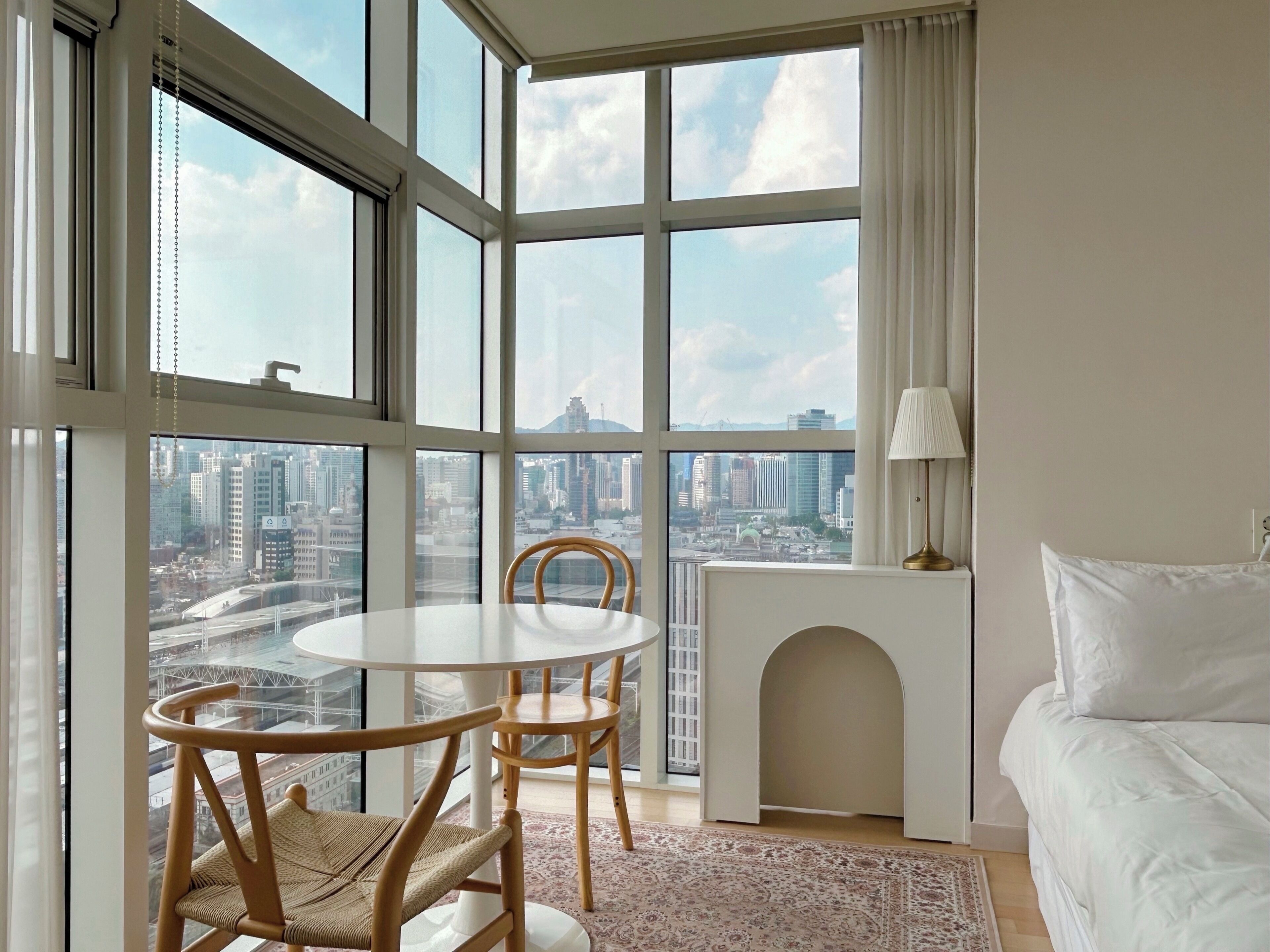 A room with a large window, a round table, two chairs, a white sofa, and a small fireplace.