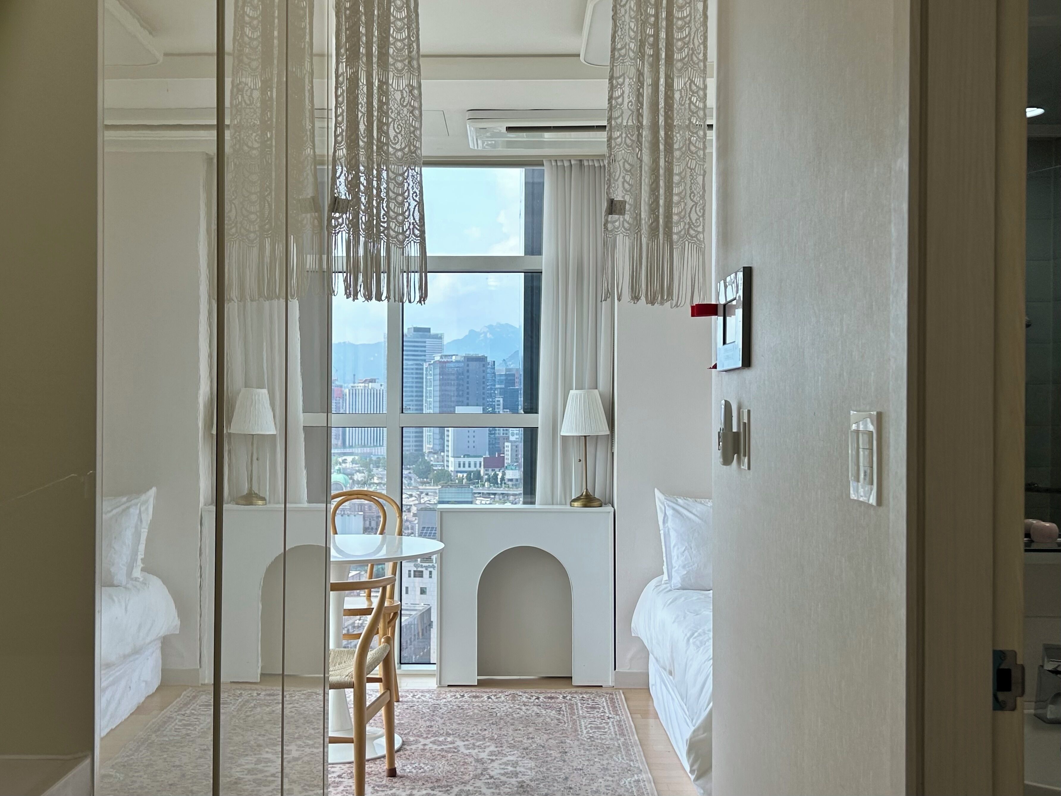 A hotel room with a bed, a desk with a chair, a window with a view of buildings, and a decorative chandelier.