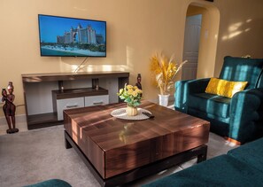 Ein modernes Wohnzimmer mit einem Holztisch, einem blaugrünen Sessel, einem Flachbildfernseher an der Wand und einer dekorativen Vase.