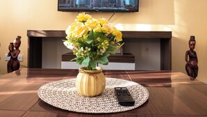 A living room with a TV displaying a cityscape, a vase with yellow flowers, and a remote control on a woven placemat. - Tulia Homestays (Nakuru)