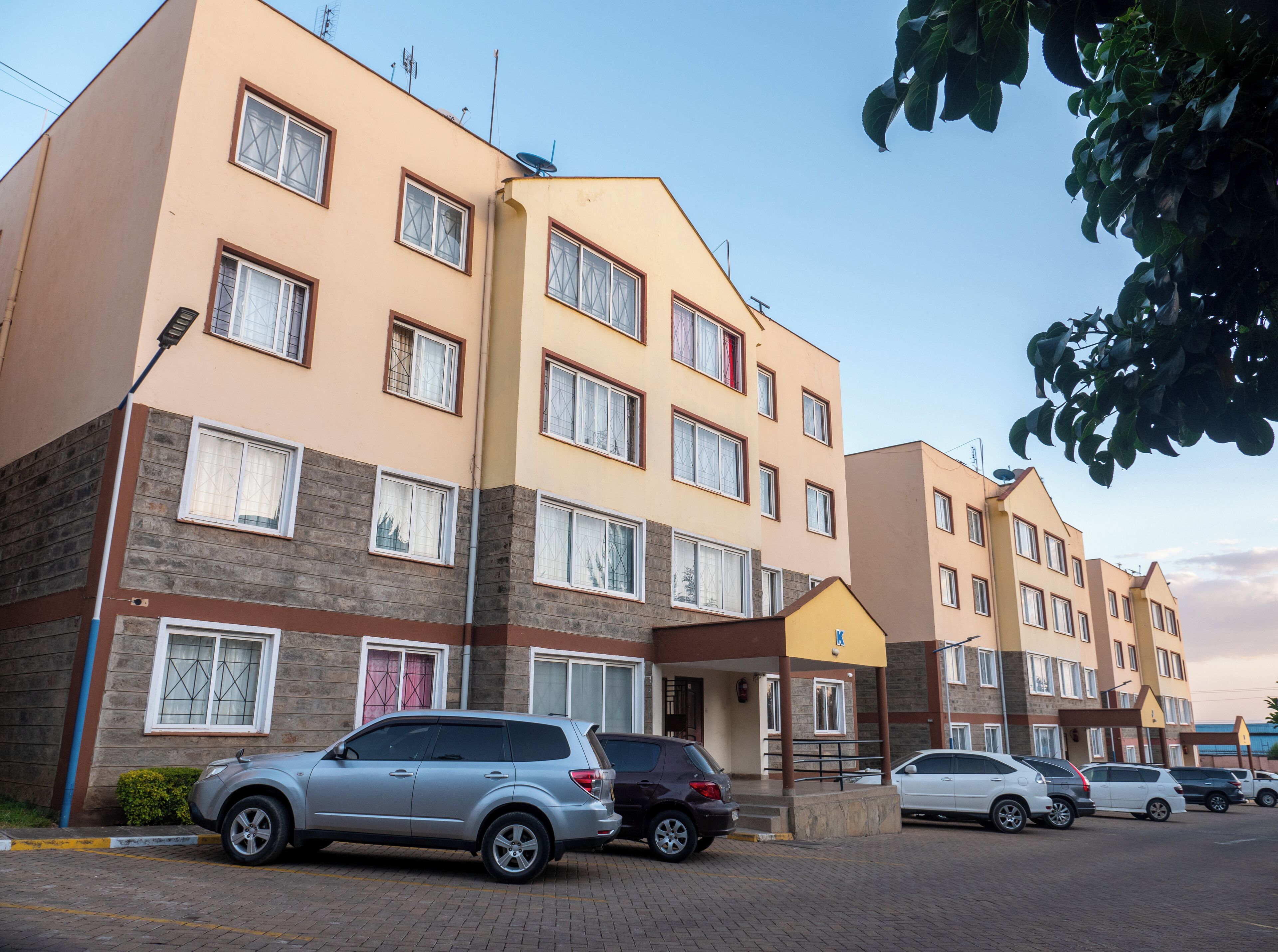 A multi-story residential building with a flat facade, multiple windows, and a parking area with several cars.