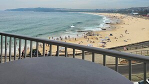 Ein Strand mit Menschen und Sonnenschirmen, eine Straße mit Verkehrsschild und eine entfernte Küstenlinie.