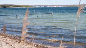 Un lago tranquilo con un muelle que se adentra en el agua, rodeado de un cielo despejado y estructuras industriales a lo lejos.