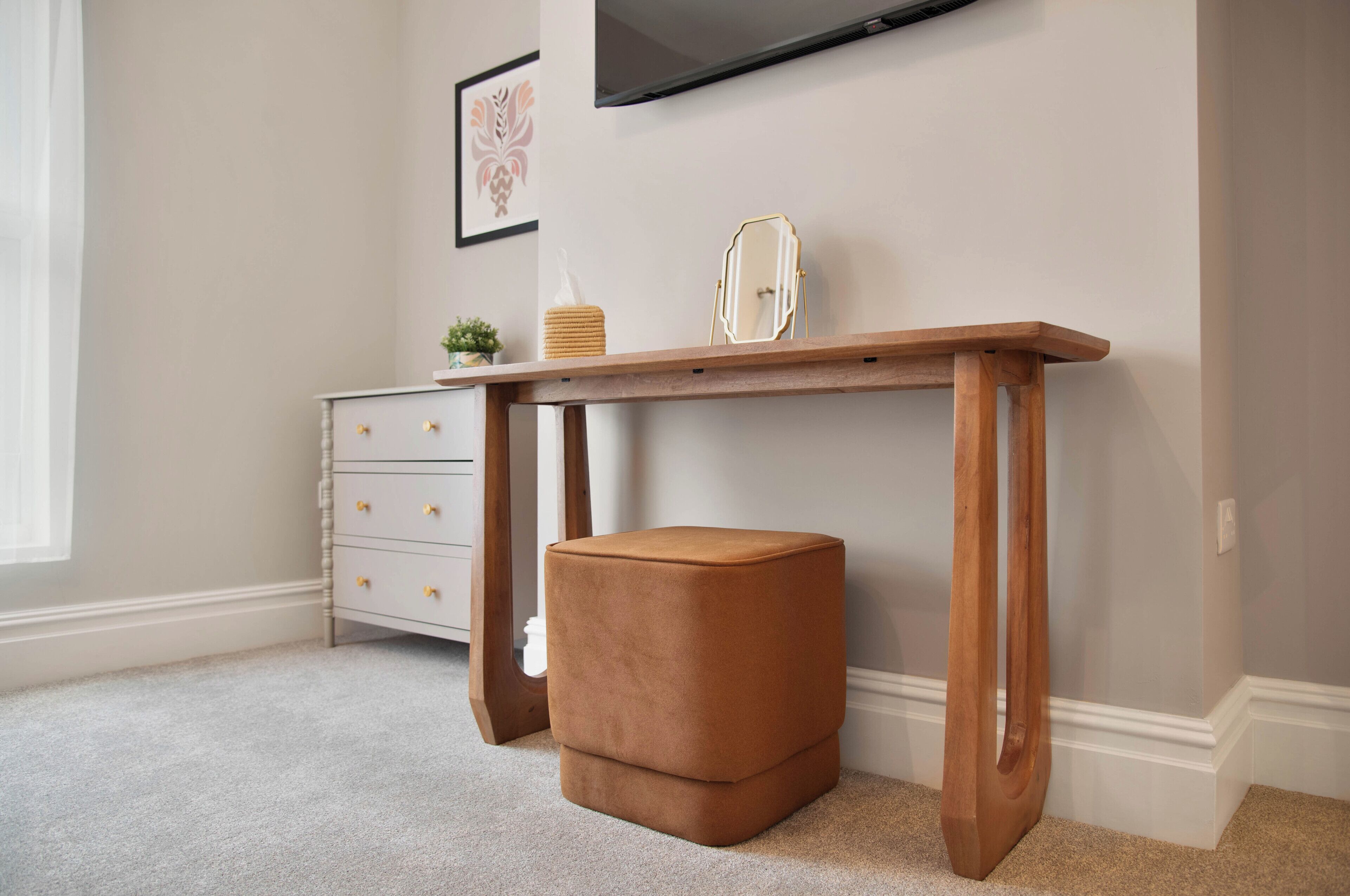 A study area with a wooden desk, a cushioned stool, and a white dresser with drawers.