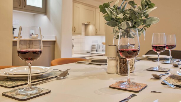 Uma mesa de jantar posta com copos de vinho, pratos e talheres, com um vaso de folhagem ao centro.