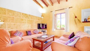 A cozy living room with a stone wall, a wooden ceiling, and a television on a stand. - Finca Roig Petit | Mallorcan Charm in Pollensa | Private Pool & Tranquil. (Pollensa)