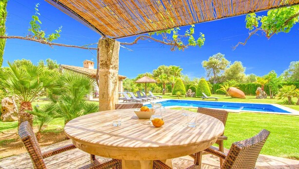 An outdoor patio with a round table and wicker chairs, a pool, and lush greenery. - Finca Roig Petit | Mallorcan Charm in Pollensa | Private Pool & Tranquil. (Pollensa)
