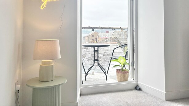 Een kamer met een neonbord, een lamp, een potplant en uitzicht op een balkon met buitenmeubilair.