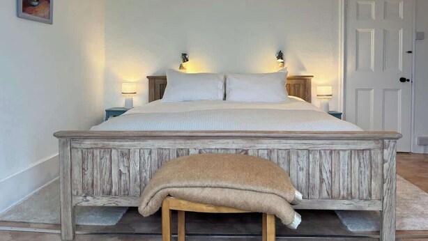 A large bed with a wooden frame and white bedding, flanked by two bedside lamps, in a room with a white door and a framed picture on the wall. - Unique Artsy Home on the Penzance Promenade (Penzance)
