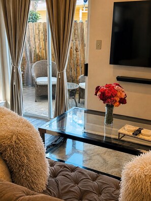 A living room with a glass coffee table, a sofa, a TV mounted on the wall, and a view of the outside. - Domain District Gem • Stylish 2BR/2BA • Perfect Location (Austin)