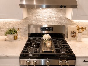 A kitchen with a stainless steel stove, a digital clock, a brick backsplash, and a book titled 'FOUR SEASONS AT HOME' on the counter. - Domain District Gem • Stylish 2BR/2BA • Perfect Location (Austin)