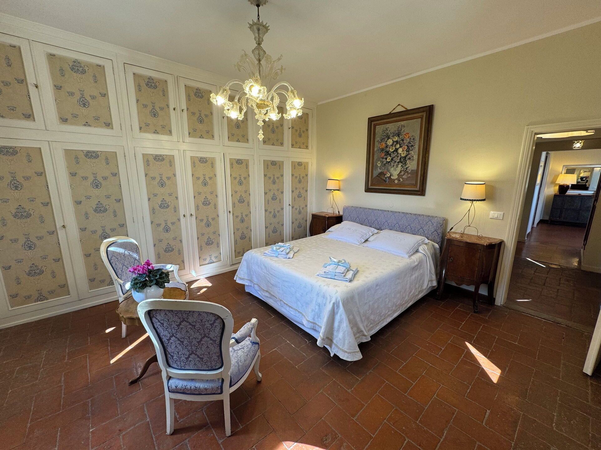 A bedroom with a large bed, two bedside tables with lamps, a chair, a chandelier, and a painting on the wall.