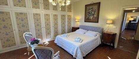 A bedroom with a large bed, two bedside tables with lamps, a chair, a chandelier, and a painting on the wall.